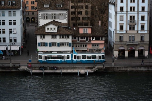 A tram goes along the water in front of some smaller houses and apartments in the city of Zurich. Photo by incusion from Pexels