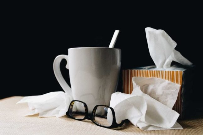 A cup of tea, tissues and Elvis Costello glasses sit next to a bed against a black wall. 